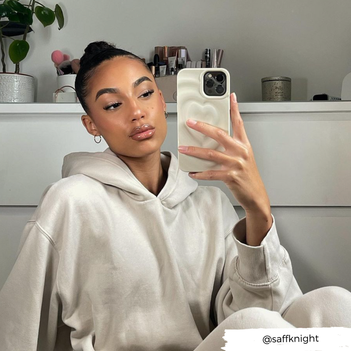 A woman with a neatly tied bun takes a mirror selfie while wearing a beige hoodie. She has a calm expression and is holding her phone, protected by the stylish Melting Heart Phone Case in Coconut Cream from Coconut Lane. The background includes a plant, books, and neatly arranged items on a white shelf. Username "@saffknight" is visible.