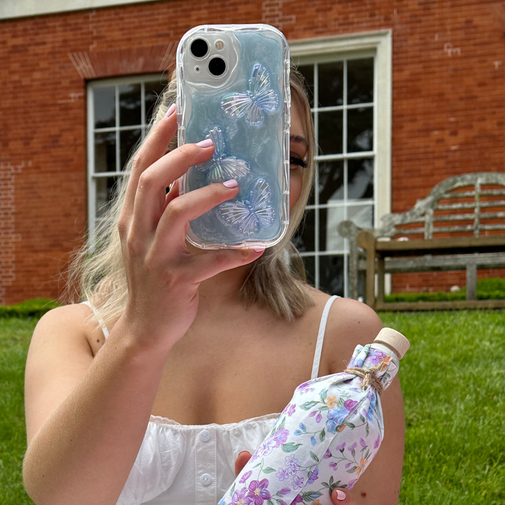A person with blonde hair, wearing a white tank top, holds up a Shimmer Blue Butterflies Phone Case by Coconut Lane, partially obscuring their face. In their other hand, they hold a floral-patterned water bottle. They are in front of a brick building and sitting on grass.