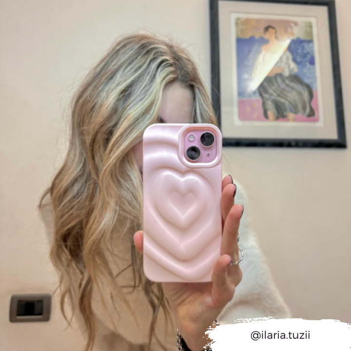 A person with long, wavy blonde hair takes a mirror selfie while holding a phone encased in the Coconut Lane Melting Heart Phone Case in Peony Pink, which features a 3D melting heart design. A piece of art is visible on the wall behind them. The Instagram handle @ilaria.tuzii is written in the bottom right corner.