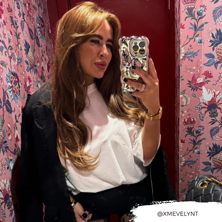 A person with long, wavy brown hair takes a mirror selfie using a Coconut Lane 3D Chrome Phone Case. They are wearing a white shirt and a watch, standing in front of a red door and floral wallpaper. Their Instagram handle "@XMEVELYNT" is overlaid at the bottom right corner.