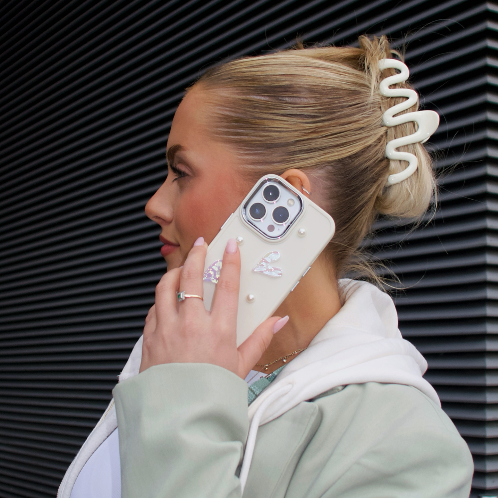 A person with blonde hair styled in a twist, adorned with a large white hair clip, holds an iPhone15 to their ear. The phone is encased in the Pearly Hearts Phone Case - Nude by Coconut Lane, featuring intricate pearl and butterfly decorations for extra protection. They are wearing a pale green and white outfit, standing against a dark striped background.