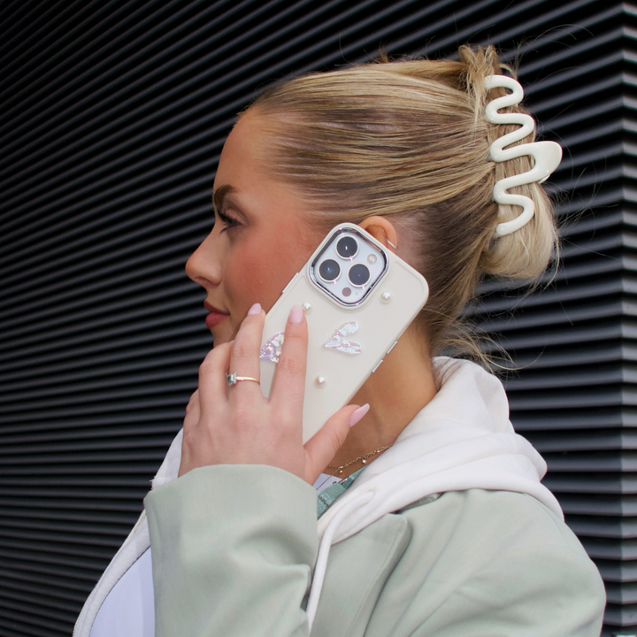 A person with blonde hair styled in a twist, adorned with a large white hair clip, holds an iPhone15 to their ear. The phone is encased in the Pearly Hearts Phone Case - Nude by Coconut Lane, featuring intricate pearl and butterfly decorations for extra protection. They are wearing a pale green and white outfit, standing against a dark striped background.