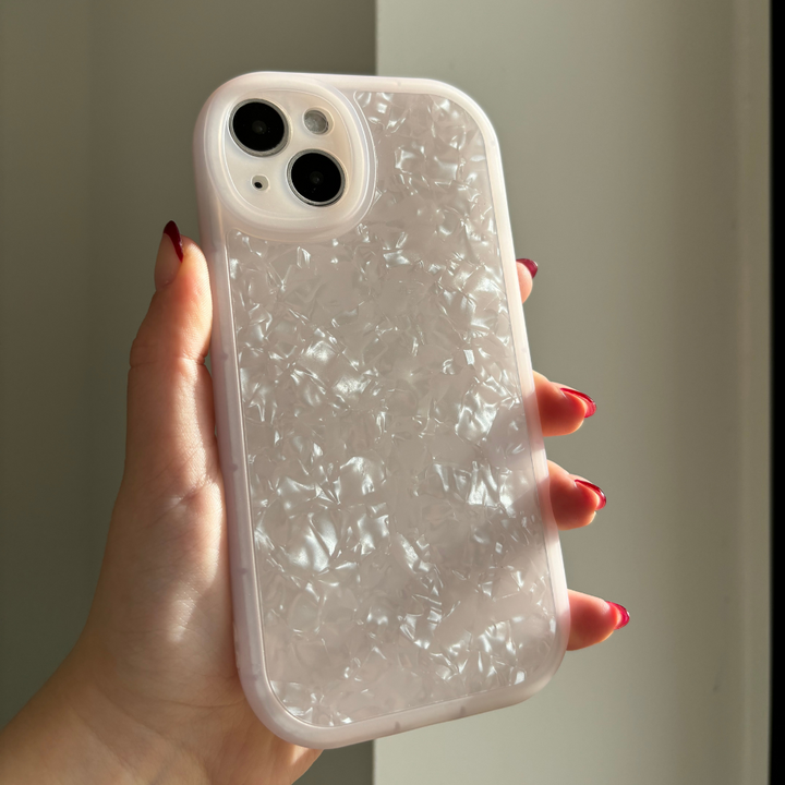 A hand with red-painted nails holds a Coconut Lane Moonstone Pink Phone Case for the iPhone15. The iridescent, protective case features a textured and translucent design that catches the light, creating a shimmering effect. The modestly lit background highlights the intricate details of the phone case.