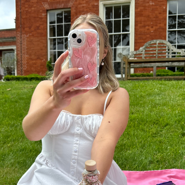 A woman in a white dress sits on a lawn in front of a brick building, holding her phone up to take a selfie. The Coconut Lane Shimmer Pink Hearts Phone Case adds an extra layer of protection while displaying glittery pink hearts. A pink picnic blanket and a small bottle are visible in the foreground.