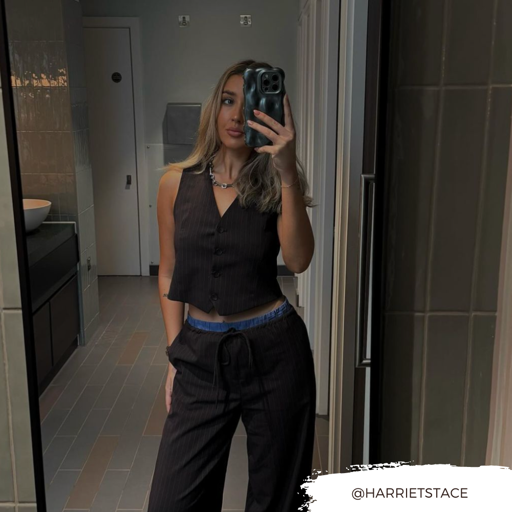 A woman with long hair takes a mirror selfie in a bathroom. She is wearing a black vest and black pants, with blue underwear slightly visible at the waistband. Her iPhone 15, encased in an extra protective 3D Pearl Phone Case - Grey by Coconut Lane, gleams in her hand. The Instagram handle "@HARRIETSTACE" is displayed in the bottom right corner.