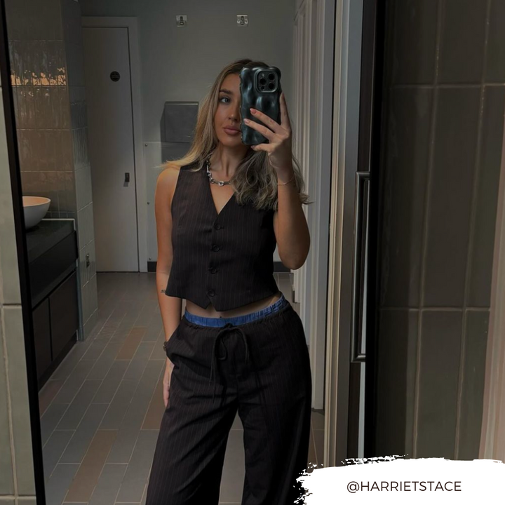 A woman with long hair takes a mirror selfie in a bathroom. She is wearing a black vest and black pants, with blue underwear slightly visible at the waistband. Her iPhone 15, encased in an extra protective 3D Pearl Phone Case - Grey by Coconut Lane, gleams in her hand. The Instagram handle "@HARRIETSTACE" is displayed in the bottom right corner.