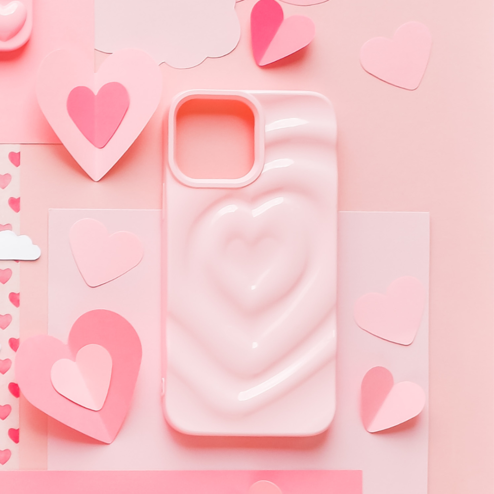 The Melting Heart Phone Case - Peony Pink by Coconut Lane is surrounded by various paper cutouts of pink and red hearts, clouds, and other decorations, all arranged on a pastel pink background.