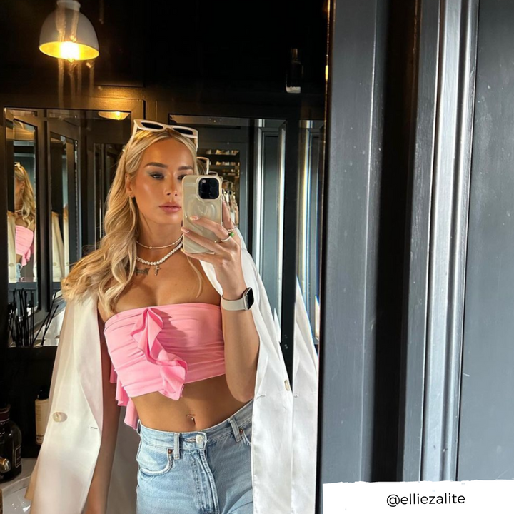 A woman with long blonde hair is taking a mirror selfie. She is wearing a pink strapless top with a bow, light denim jeans, and a white blazer draped over her shoulders. Sunglasses rest on her head as she holds her phone in its Melting Heart Phone Case - Coconut Cream by Coconut Lane. The Instagram handle @elliezalite is displayed in the bottom right corner.