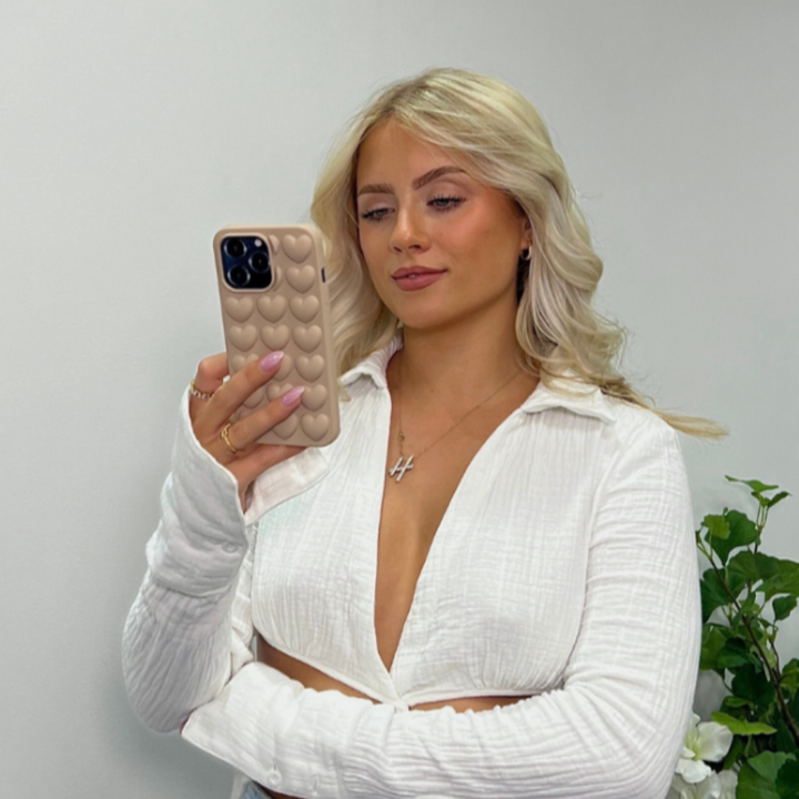A person with long blonde hair takes a selfie in front of a light-colored wall, wearing a white, buttoned, long-sleeve top with an open collar and a cross necklace. Their iPhone 15 is encased in Coconut Lane's 3D Hearts Phone Case - Matte Nude. Some green plants are visible in the background.