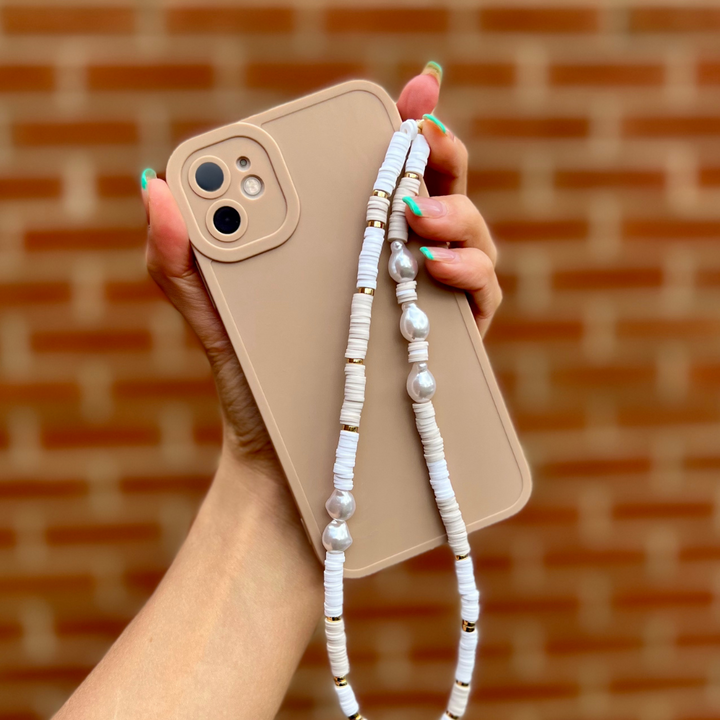 A hand with green nail polish holds a beige smartphone adorned with the chic Coconut Lane Beaded Phone Strap - Nude & Pearl. The strap features white and gold beads, interspersed with a few larger silver ones. A brick wall is blurred in the background, enhancing the stylish scene.