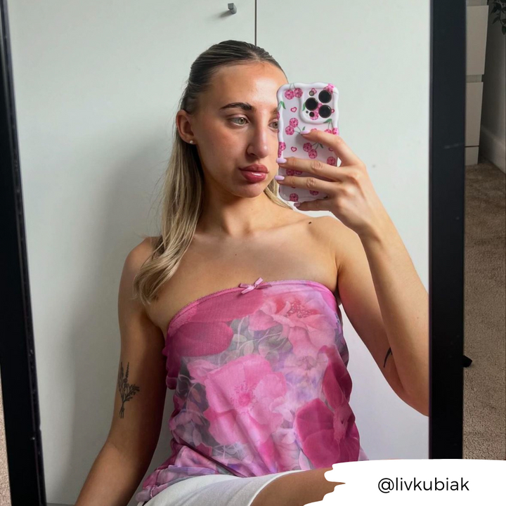 A person with long blonde hair takes a mirror selfie. They are wearing a pink strapless top with a floral pattern and hold a phone featuring Coconut Lane's extra protective Wavy Phone Case - Disco Cherries. A tattoo is visible on their right forearm, and the username "@livkubiak" appears in the bottom right corner.