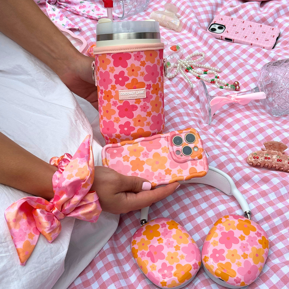 A person is holding the Coconut Lane Wavy Phone Case - Pink & Orange Bloom, seated on a pink checkered picnic blanket. The scene features matching floral accessories like a scrunchie, drink holder, headphones, and various items including a pearl necklace and eyeglasses, all in a coordinating pink theme.