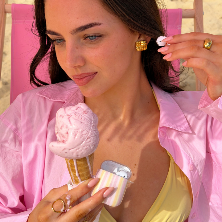 Girl putting airpods in her ear holding ice cream on the beach with her airpods case pastel stripes