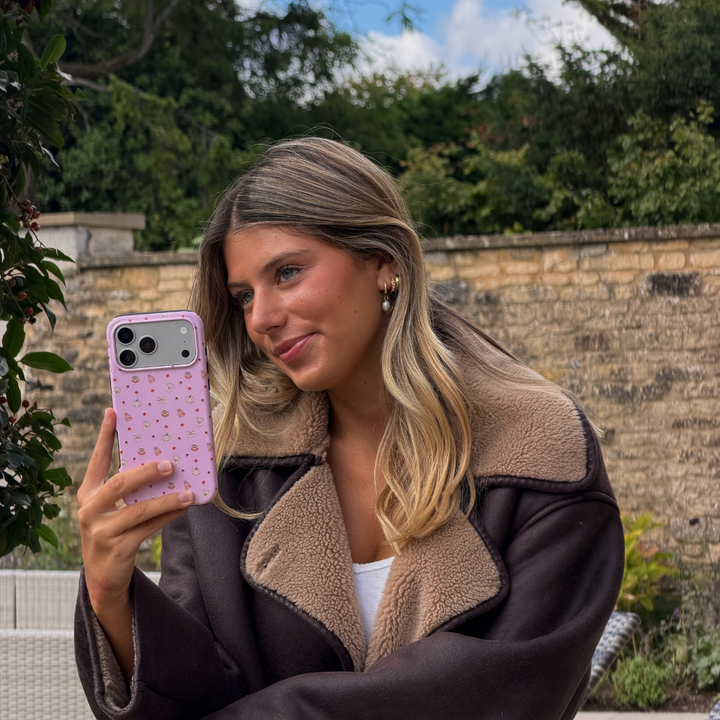 Woman wearing brown aviator jacket holds up an iphone taking a photo, in a baby pink phone case, with small dainty pastries, pumpkins, wooly hats, bows and leaves printed across the case.