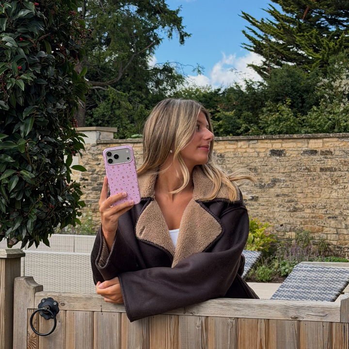 Woman wearing brown aviator jacket holds up an iphone , whilst leaning over a wooden gate, showing a baby pink phone case, with small dainty pastries, pumpkins, wooly hats, bows and leaves printed across the case.