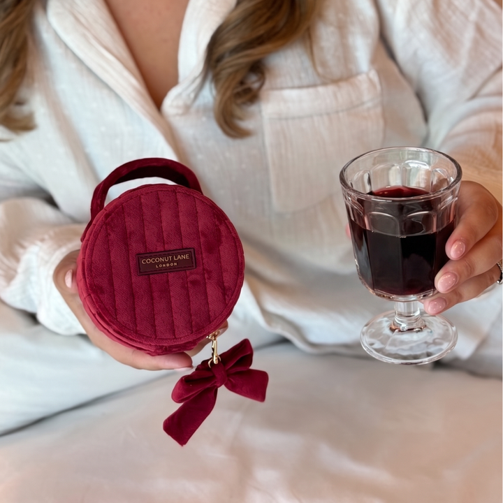 Burgundy velvet jewellery box with a quilted design, top handle, bow detail, and gold-tone hardware. Girl is holding it with a glass of red wine
