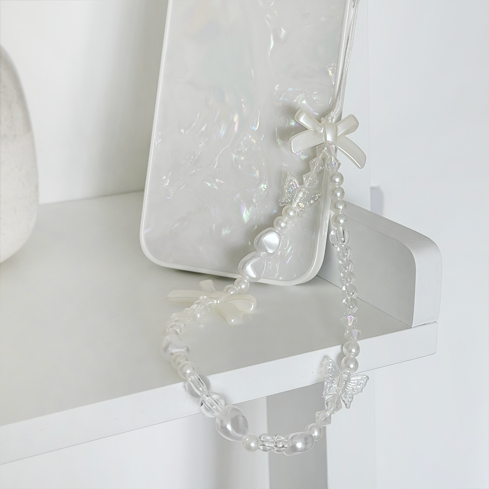 A Beaded Phone Strap - Bows & Butterflies by Coconut Lane, featuring charms of bows and butterflies, hangs from a pearlescent phone case placed on a white surface. The phone accessory leans against a white wall, creating a minimalist and elegant setting.