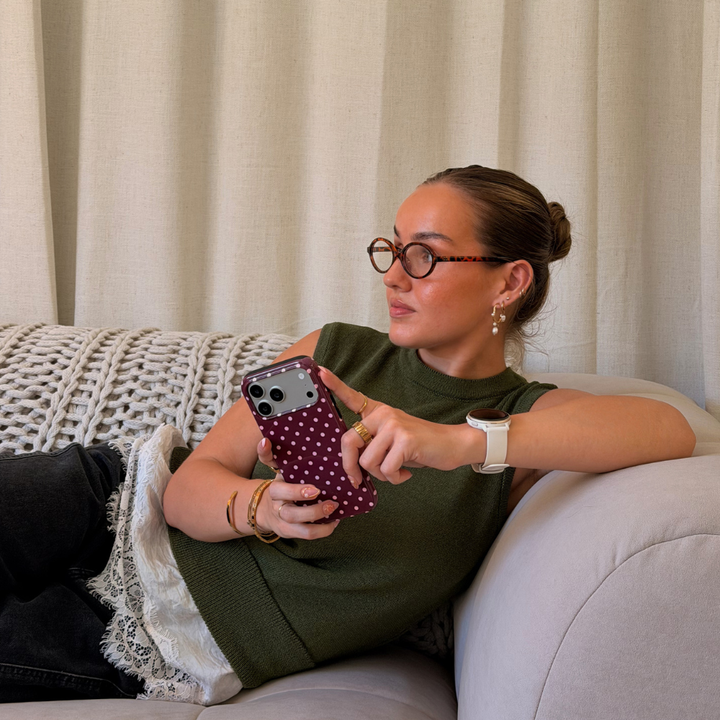 Woman wearing a dark green jumper with a white lace shirt underneath , lace on a beige sofa. She is holding an iphone in a browny burgundy case with baby pink polka dots. 