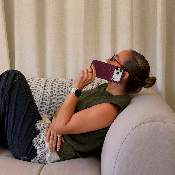 Woman wearing a dark green jumper with a white lace shirt underneath , lace on a beige sofa. She is holding an iphone up to her ear, the phone is in a browny burgundy case with baby pink polka dots. 