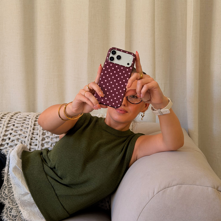Woman wearing a dark green jumper with a white lace shirt underneath , lace on a beige sofa. She is holding an iphone in a browny burgundy case with baby pink polka dots. 