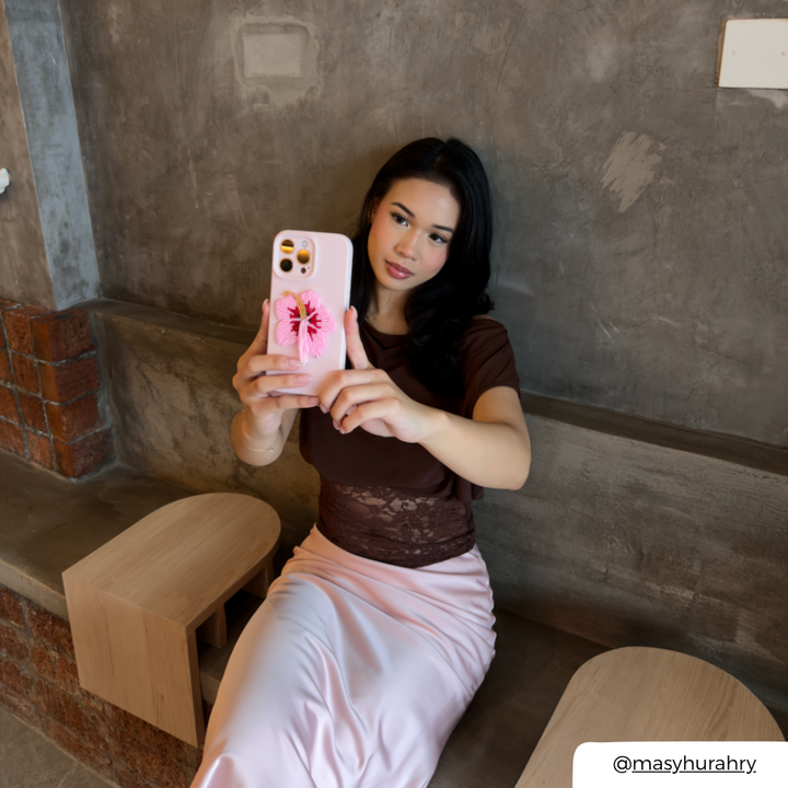 A woman taking a mirror selfie in a cafe, holding a light pink phone case decorated with a pink and red flower beaded phone patch. She is wearing a brown lace-trimmed top and a light pink satin skirt, seated on a wooden bench against a textured concrete wall.