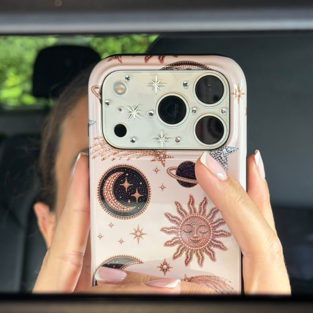 A close-up of a phone case held in front of a car mirror, showing a celestial pattern with a sun, moon, stars, and planets in rose gold and black on a pale pink background. A person's hands and face are visible holding the phone.