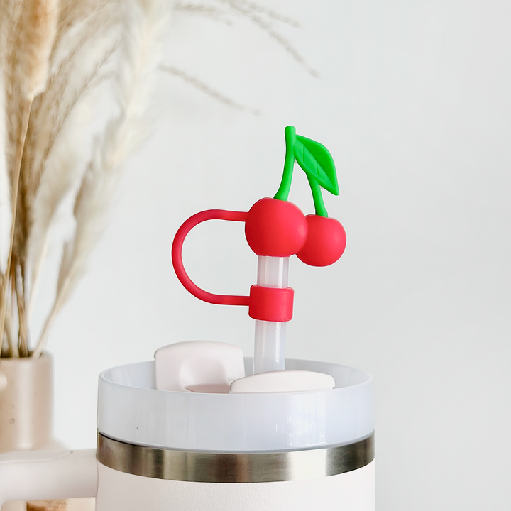 The Cherries Straw Cover by Coconut Lane features a white tumbler adorned with a silver rim, complemented by a uniquely designed silicone straw topped with a red cherry-shaped decoration and green leaves for added durability. The cover ensures the straw remains hygienic, while in the background, a vase filled with dried pampas grass enhances the minimalist and clean aesthetic.