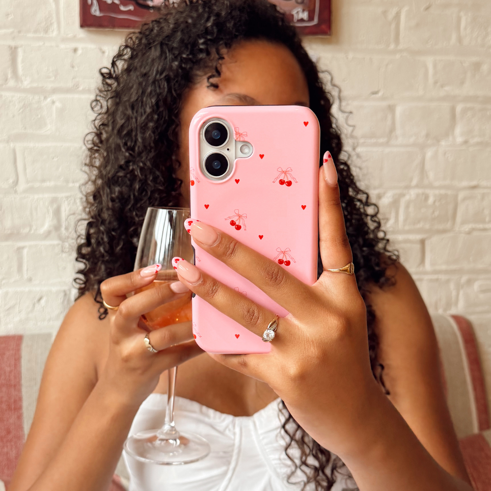 Girl holding a pink phone with heart and cherry design and a glass of wine.