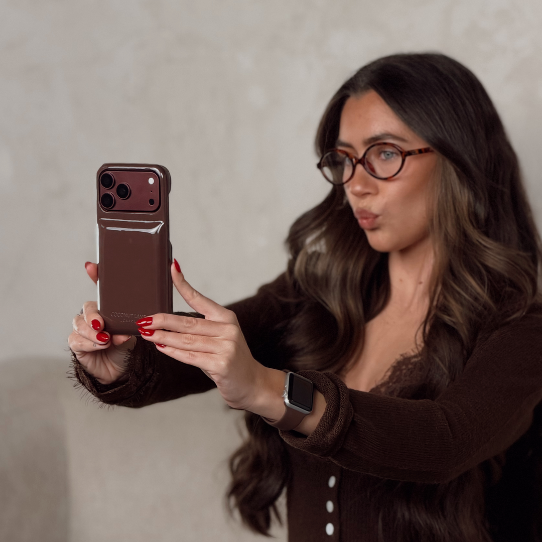 Woman wearing brown cardigan with lace detail, taking a selfie, holding a Glossy chocolate brown Coconut Lane puffer phone case with embossed logo. On her wrist is a Coconut Lane NAKD Chocolate brown apple watch strap.