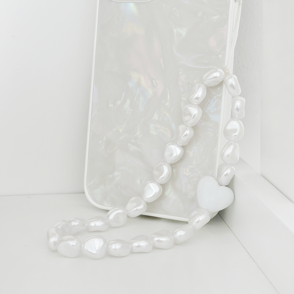 A white, iridescent phone case is adorned with Coconut Lane's Beaded Phone Strap - Chunky Pearls, featuring a small white heart charm. The phone case is propped up and leaning against a white surface, creating a striking contrast with the glossy and shiny appearance of the case and strap.