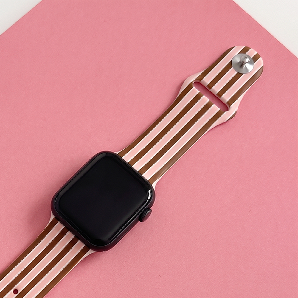 Close-up of the Cocoa & Pink Stripe Apple Watch strap, showing the striped silicone design, silver fastening pin and black watch face against a pink background.