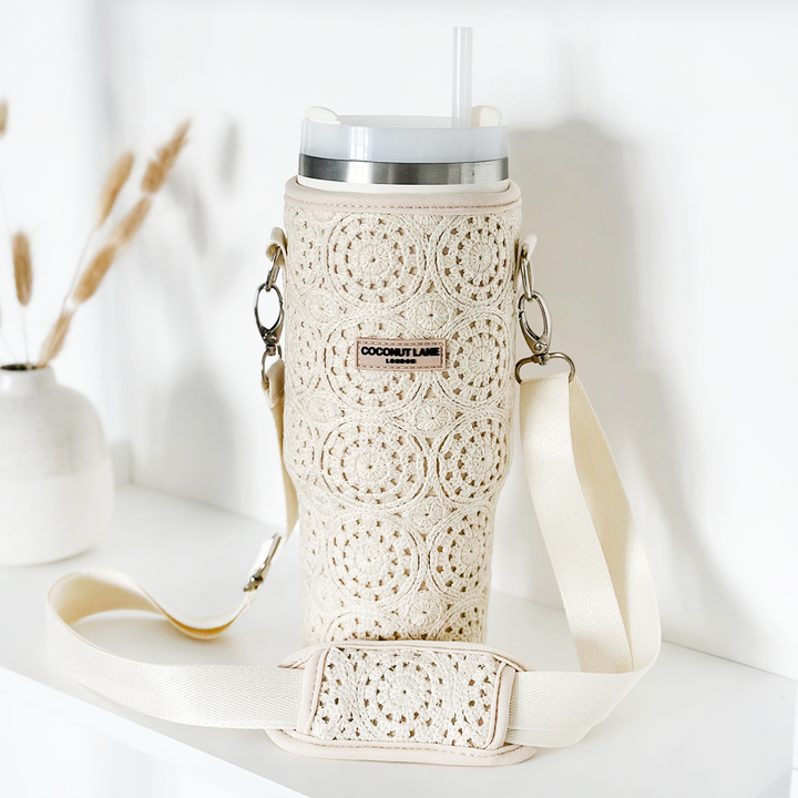 A beige Tumbler Carry Case - Festival Crochet by Coconut Lane with a shoulder strap is shown on a white surface. The case houses a metal cup with a lid and straw inside, ensuring optimal tumbler protection. In the background, there is a white vase with dried grass stems.