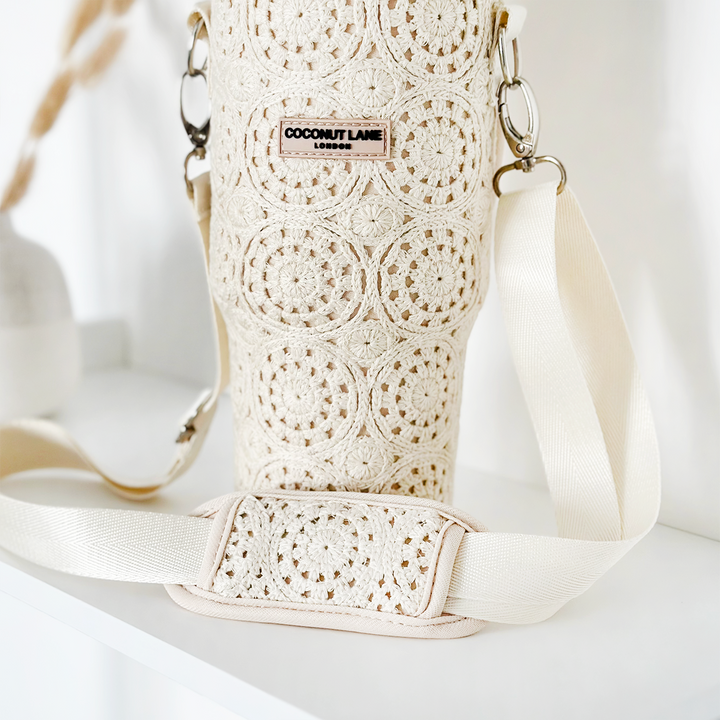 The Tumbler Carry Case - Festival Crochet by Coconut Lane is displayed. This beige crochet water bottle holder showcases intricate circular patterns and includes a cream-colored adjustable strap with a padded section for added comfort. The travel accessory features a branded label reading "COCONUT LANE LONDON." It is placed on a white surface.