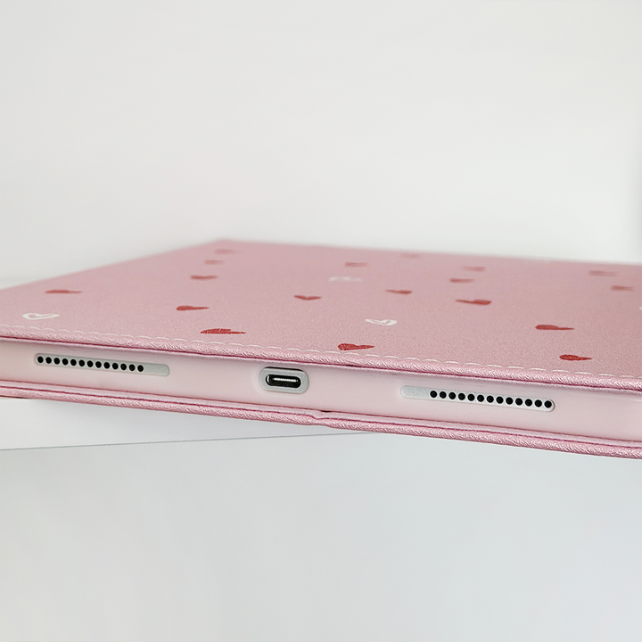 Close-up of the Cute Hearts iPad Case by Coconut Lane, crafted from PU vegan leather and adorned with small red and white hearts. The image highlights the bottom edge of the case, showcasing a central charging port flanked by two speaker grilles. The background is plain white.