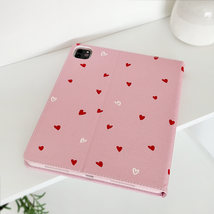 A pink Coconut Lane Cute Hearts iPad Case, adorned with a charming pattern of small red and white hearts, is displayed on a white surface. The open camera slot clearly suggests compatibility with a device that has a rear camera. A hint of a plant can be seen in the lower left corner, accompanied by a white ceramic vase in the upper right corner.