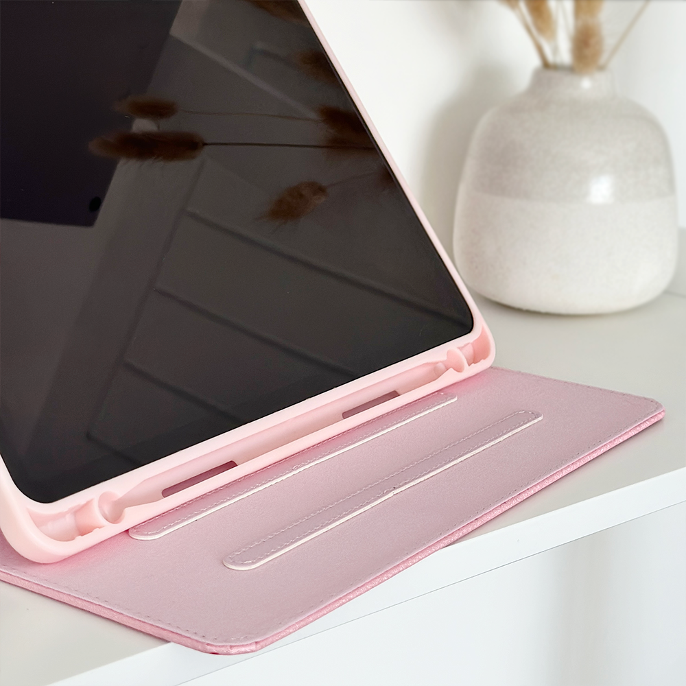 A tablet encased in Coconut Lane's pink Cute Hearts iPad Case is propped up on a white shelf, beside a white vase filled with dry ornamental grasses. The screen of the tablet is turned off, reflecting part of the surroundings. The overall scene features a soft, minimalistic aesthetic.