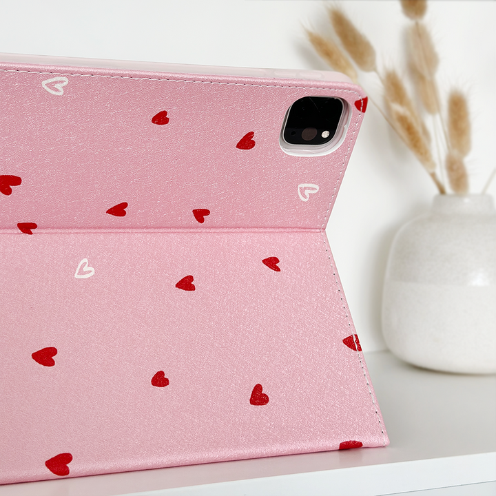 The Cute Hearts iPad Case by Coconut Lane is an adorable cover made from PU vegan leather, featuring a pink design adorned with small red and white hearts. It rests on a white surface, complemented by a white ceramic vase filled with dried wheat stalks for a natural touch.
