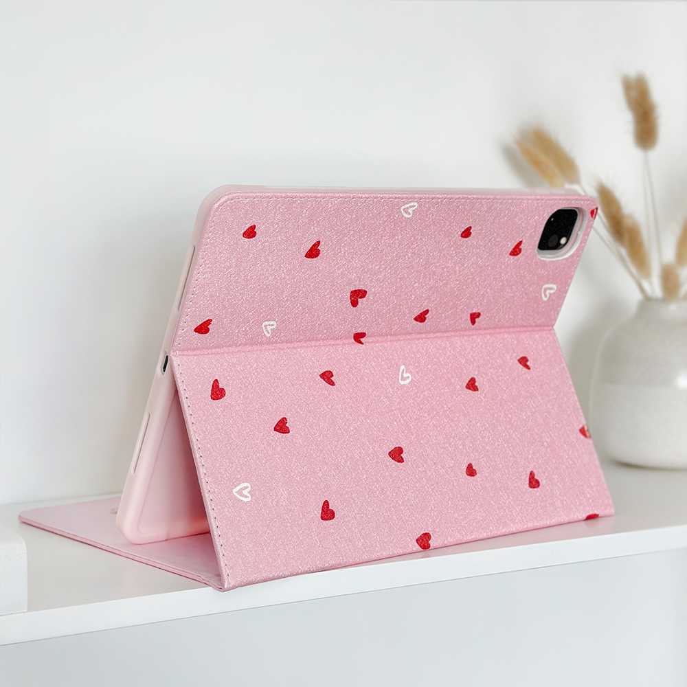 A pink Cute Hearts iPad Case by Coconut Lane, crafted from PU vegan leather, sits open on a white shelf. The case, adorned with a charming red and white heart pattern, holds an iPad. In the background, a white vase filled with dried beige grasses enhances the minimalist scene with a touch of natural decor.