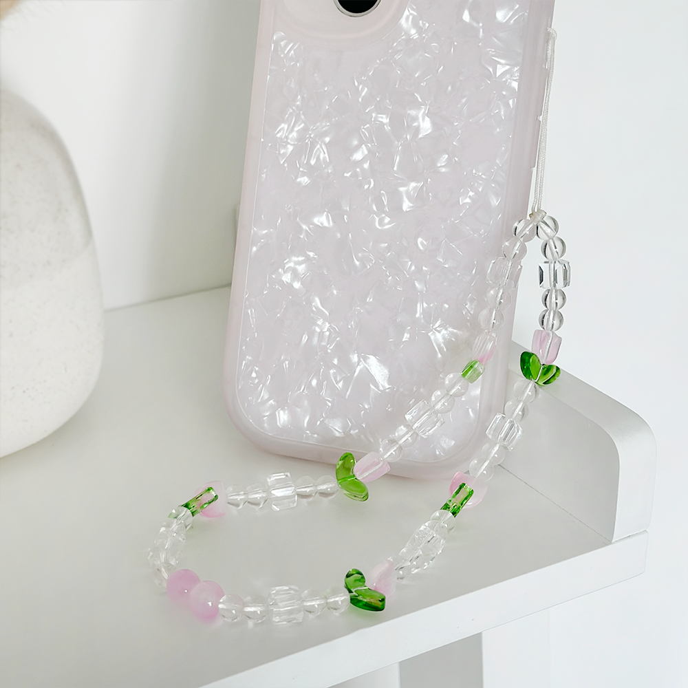 A smartphone with a textured, pearlescent white case is placed on a white surface. Attached to the phone is the "Beaded Phone Strap - Cute Tulips" by Coconut Lane, made of clear, pink, and green beads, which rests neatly on the same surface. A small portion of a plant pot is visible in the corner.