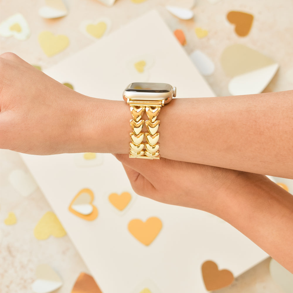 A person's wrist adorned with a Coconut Lane gold smartwatch featuring the Hearts Chain Apple Watch Strap. The background includes heart-shaped confetti in various shades of gold and white scattered around, creating a festive and elegant atmosphere.