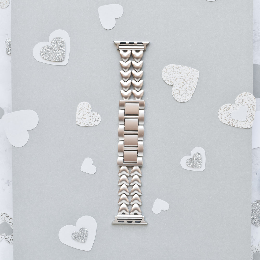 A Starlight Hearts Chain Apple Watch Strap by Coconut Lane, featuring heart-shaped links in a silver hue, is displayed on a light gray background. Small silver and white paper hearts are scattered around, creating a romantic and elegant theme. This stainless steel adjustable strap fits ALL Apple Watch models perfectly.