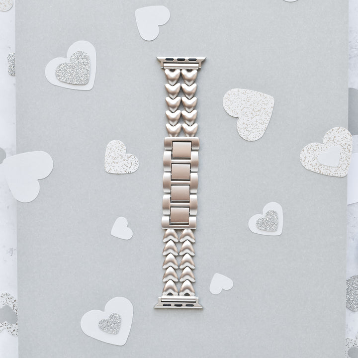 A Starlight Hearts Chain Apple Watch Strap by Coconut Lane, featuring heart-shaped links in a silver hue, is displayed on a light gray background. Small silver and white paper hearts are scattered around, creating a romantic and elegant theme. This stainless steel adjustable strap fits ALL Apple Watch models perfectly.