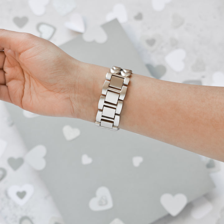 A person is wearing a Hearts Chain Apple Watch Strap - Starlight from Coconut Lane on their wrist. The background features a gray surface scattered with white and silver heart shapes, accentuating the watch strap that fits ALL Apple Watch models.