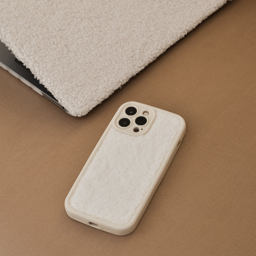 A cream-colored phone with a soft, fuzzy Cosy Teddy Phone Case by Coconut Lane is resting on a light brown surface. Partially visible beside it is a matching extra-protective fuzzy laptop cover. The cozy, soft texture of these items by Coconut Lane stands out.