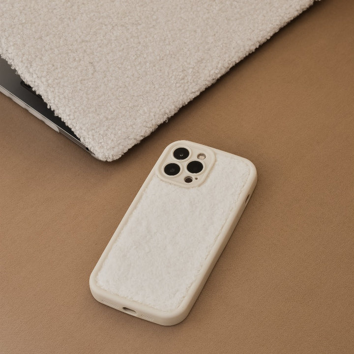 A cream-colored phone with a soft, fuzzy Cosy Teddy Phone Case by Coconut Lane is resting on a light brown surface. Partially visible beside it is a matching extra-protective fuzzy laptop cover. The cozy, soft texture of these items by Coconut Lane stands out.