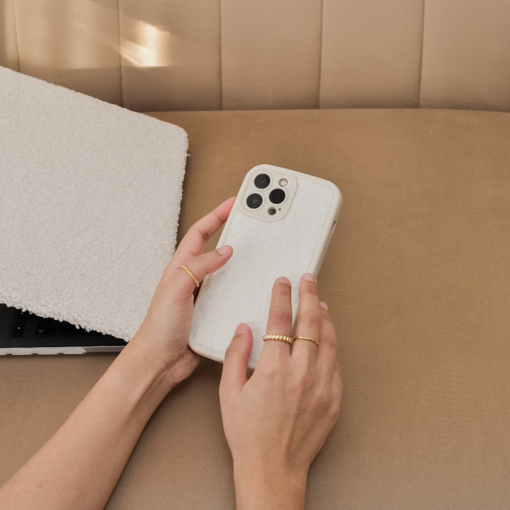 A person with manicured hands is holding a smartphone encased in Coconut Lane's Cosy Teddy Phone Case - Cream, known for its extra protective, textured teddy finish. The phone hovers above a beige leather-like surface, partially covering a laptop adorned with a similarly textured cover. In the background, a quilted fabric serves as the backdrop.