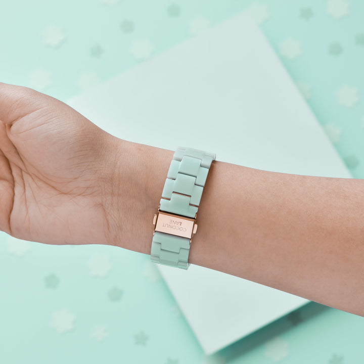 A hand wearing a Coconut Lane Luxe Pastel Mint Apple Watch Strap, featuring a rose gold clasp, is set against a mint green background adorned with small white floral shapes. The watch has an adjustable, linked strap, creating a harmonious and aesthetically pleasing scene.