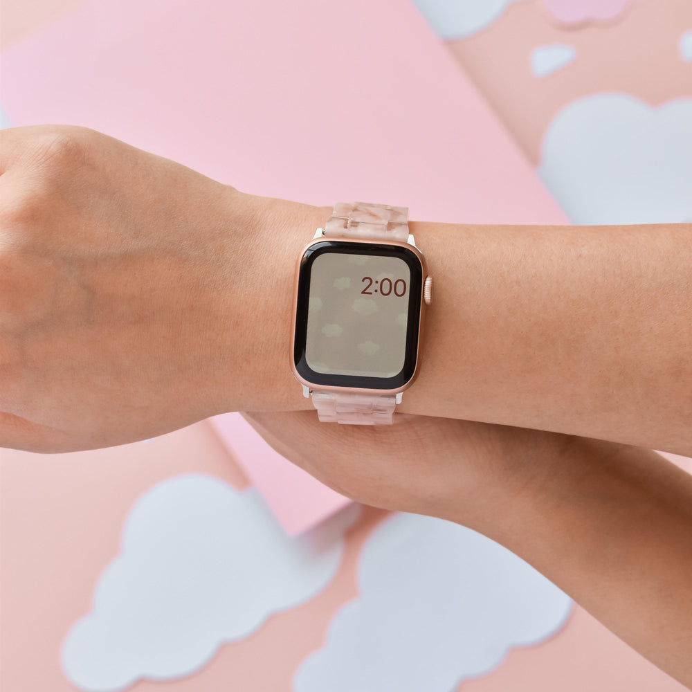A person wearing the Luxe Pink Clouds Apple Watch Strap by Coconut Lane shows the time as 2:00. The background consists of pastel pink and white cloud-like shapes. Only the wrist and hand of the person are visible, with the smartwatch prominently displayed.