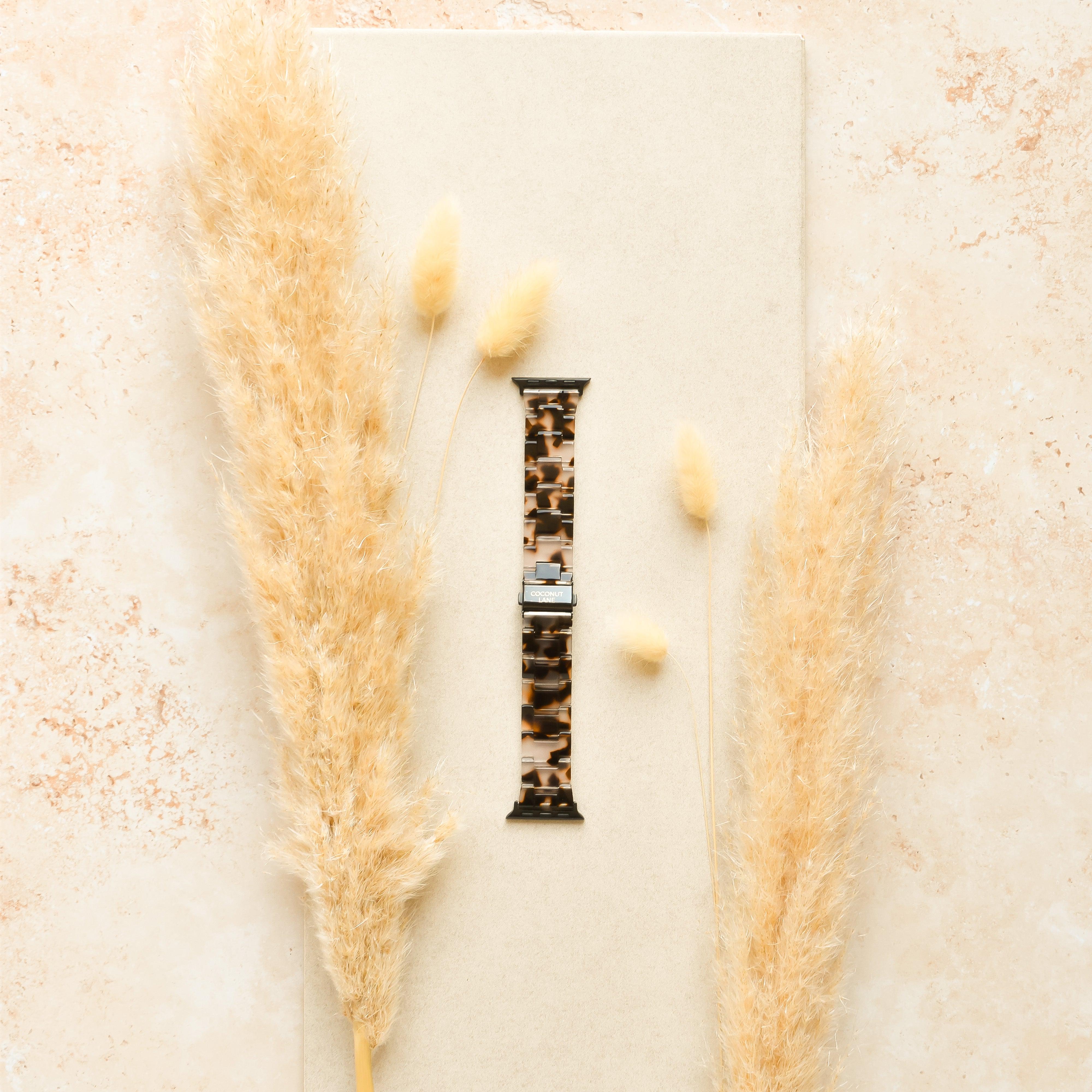 A Luxe Ivory Tort Apple Watch Strap by Coconut Lane is placed on a beige surface, surrounded by dried pampas grass. The background features a matching beige-brown hue, creating a warm and natural aesthetic.