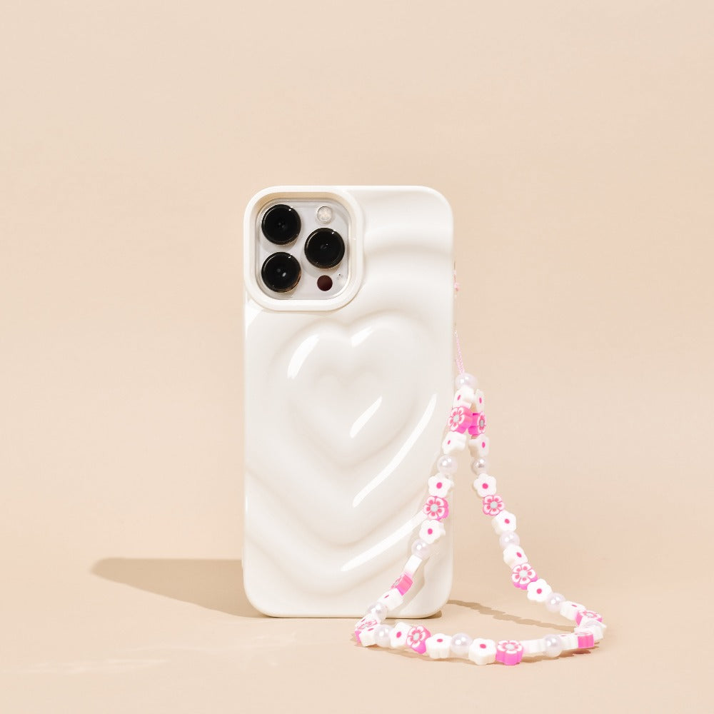 A white smartphone with a heart-shaped design on its back cover stands upright against a beige background. Attached is the Beaded Phone Strap - Flowers & Pearls by Coconut Lane, featuring pink and white beads, including charming flower-shaped ones, making it the perfect decorative accessory.